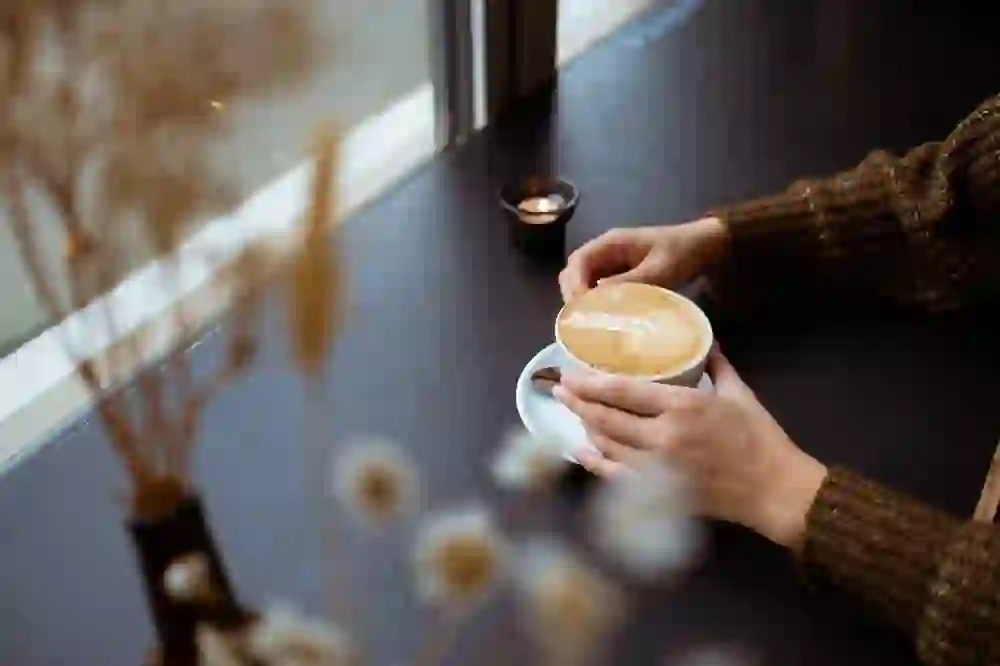 Auf dem Foto sind die Hände einer Person zu sehen, die eine Tasse Cappuccino halten, auf dessen Schaum ein Latte-Art-Blattmuster zu erkennen ist. Der Tisch, an dem die Person sitzt, steht nahe einem Fenster, durch das man verschwommen die Außenwelt sieht. Im Hintergrund ist ein Teelicht zu erkennen. Die Atmosphäre wirkt gemütlich und entspannt.