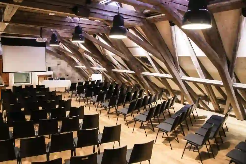 The picture shows a spacious attic seminar room with exposed, rustic wooden beams, complemented by modern black chairs and lighting - an impressive fusion of old and new.