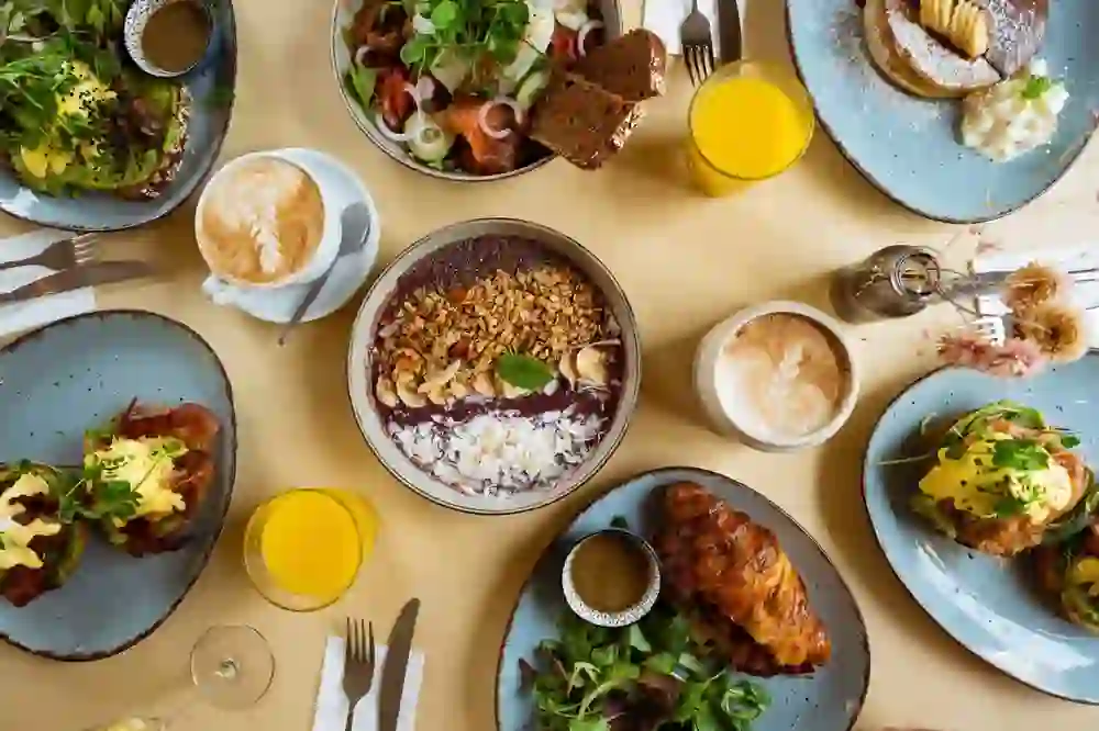 The picture shows a varied brunch menu on a table. You can see an acai bowl with nuts, a plate of scrambled eggs and avocado on toast, a croissant next to a salad, pancakes with icing sugar and side dishes, as well as a salmon platter with dark bread. There are also drinks such as orange juice, cappuccino and a glass of water.