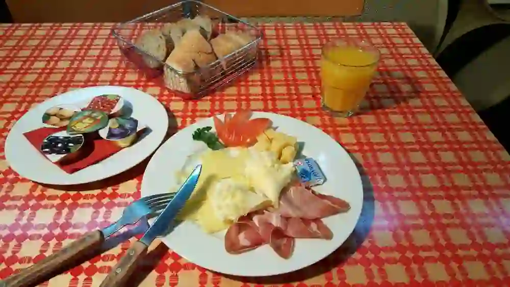 L'immagine mostra un tavolo per la colazione nel ristorante Le Lötschberg AOC, apparecchiato con vari piatti. Tra questi, diversi tipi di formaggio, prosciutto, un bicchiere di succo d'arancia e un cestino con diversi tipi di pane. La tovaglia ha un motivo rosso.