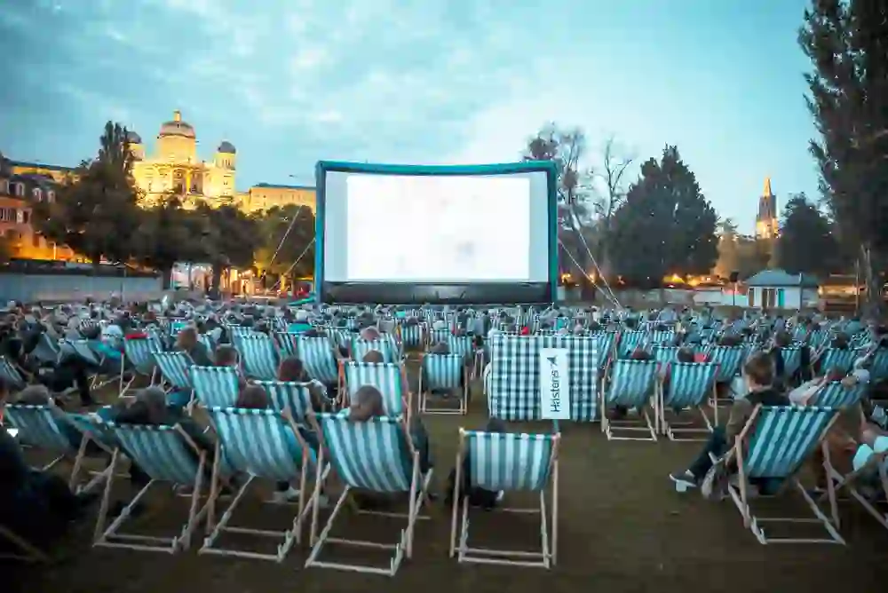 Un cinema all'aperto a Berna: il crepuscolo illumina l'architettura storica sullo sfondo, mentre una folla su sedie a sdraio a righe guarda con attenzione il grande schermo dell'Opernairkino Marzili. L'atmosfera è accogliente e comunitaria, come se tutta la città si riunisse per godere della magia del cinema sotto le stelle.
