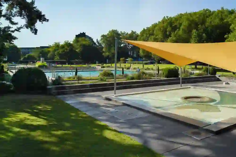 L'immagine mostra un angolo della piscina all'aperto Wyler di Berna, un rifugio soleggiato e tranquillo. Una piccola vasca per bambini in primo piano e una grande piscina sullo sfondo, circondate dal verde e da alberi ombrosi, invitano a soffermarsi.