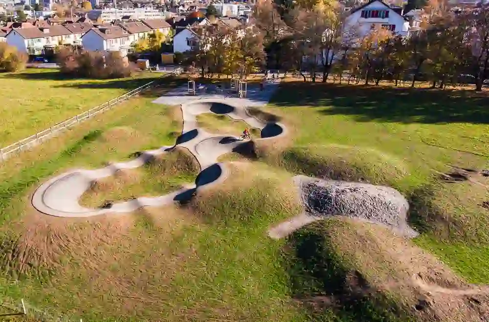 From a bird's eye view, we see a pump track in Ostermundigen, artistically designed and surrounded by an urban landscape. The curved lanes of the track are a playground for cyclists, who are clearly enjoying themselves there, embedded in an environment that harmoniously combines urban life and active leisure sport.