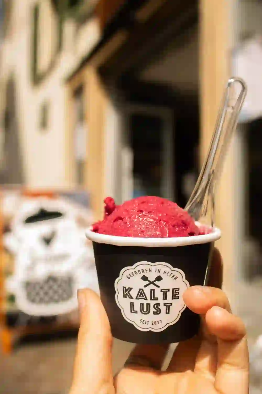 L'immagine cattura il piacere dell'estate: una mano regge una tazza di sorbetto fruttato di "Kalte Lust" su uno sfondo urbano sfocato - un momento di piccola felicità.