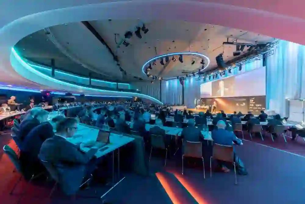 Das Bild zeigt eine Konferenzhalle mit Menschen, die an Tischen sitzen und auf eine Bühne schauen, wo eine Person spricht. Große Bildschirme zeigen den Sprecher. Die Beleuchtung ist blau und rot.