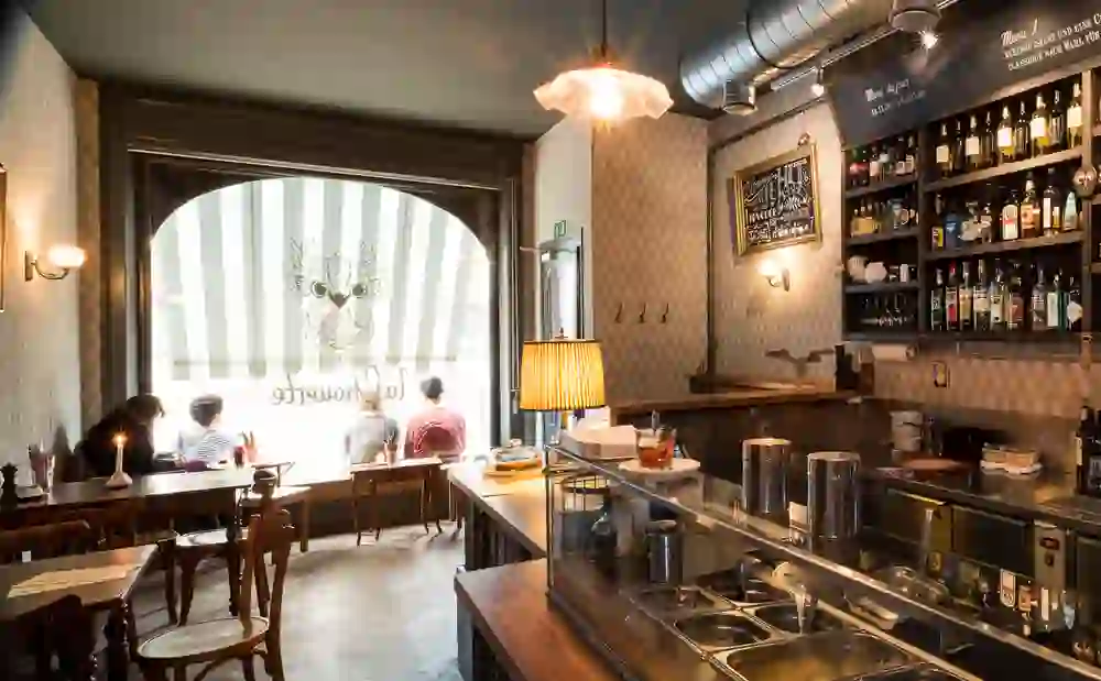 Das Bild zeigt das gemütliche Innere eines Cafés mit warmem Licht und eleganter Einrichtung. Gäste sitzen entspannt am Fenster, genießen Getränke und die ruhige, einladende Atmosphäre. Die Dekoration vermittelt einen charmanten, altmodischen Flair.