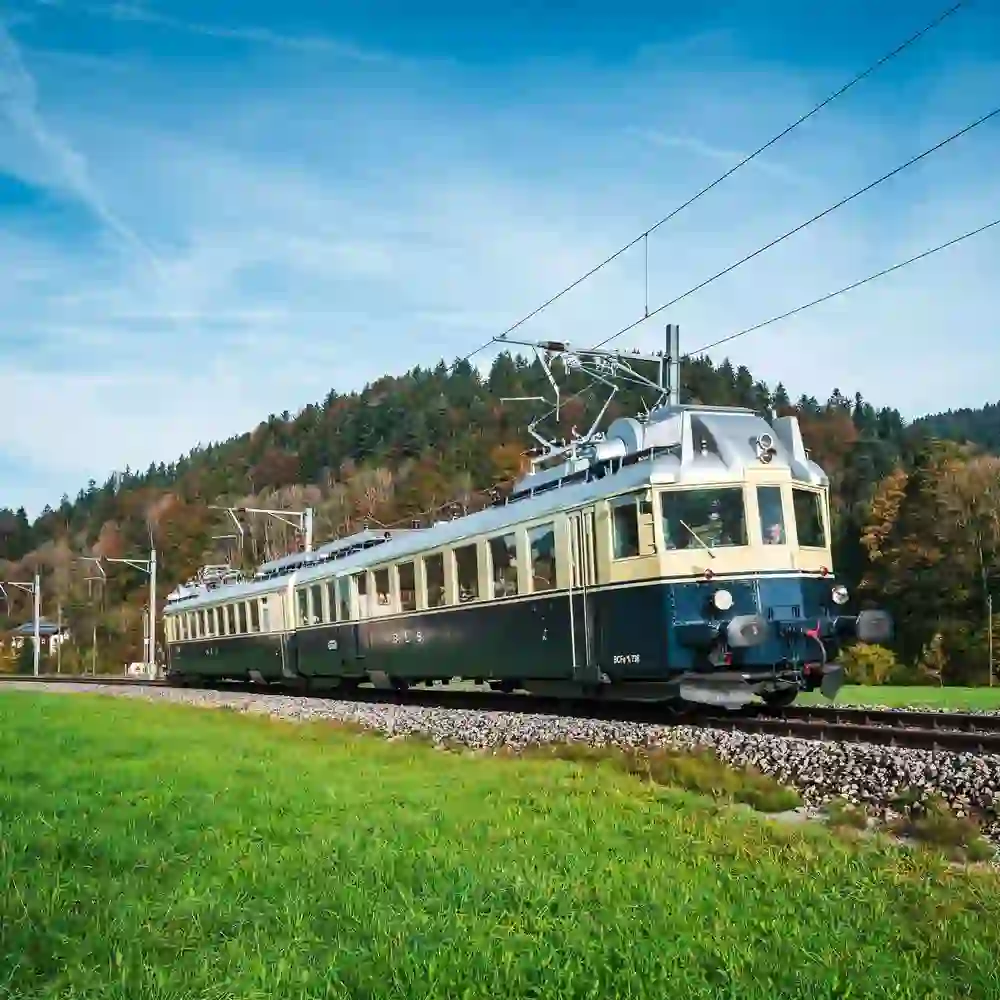 Un nostalgico treno della birra BLS verde e beige attraversa un paesaggio idilliaco. L'atmosfera soleggiata e la vegetazione lussureggiante trasmettono una sensazione di pace e piacere, come un invito a fare un accogliente viaggio nel tempo.