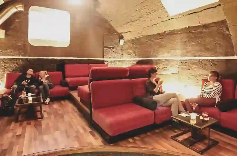 Das Foto zeigt die gemütliche Abflugbar mit Steinmauern und weichen, roten Sofas, die zum Entspannen einladen. Zwei Personen sitzen lässig und unterhalten sich bei einem Getränk. Die Atmosphäre ist warm und einladend, wie in einer schicken Lounge.