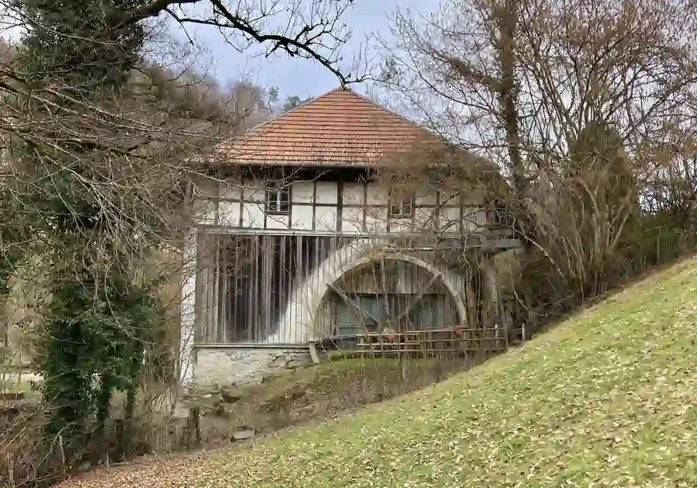 Historic half-timbered house with an arched entrance, surrounded by bare trees and a grassy hill, under a cloudy sky.