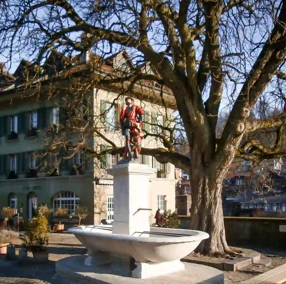 Das Bild zeigt den Läuferbrunnen in Bern, Schweiz. Es ist ein historischer Brunnen, der einen Läufer in traditioneller Kleidung darstellt. Der Brunnen steht auf einem Platz, umgeben von Bäumen und einem historischen Gebäude im Hintergrund.