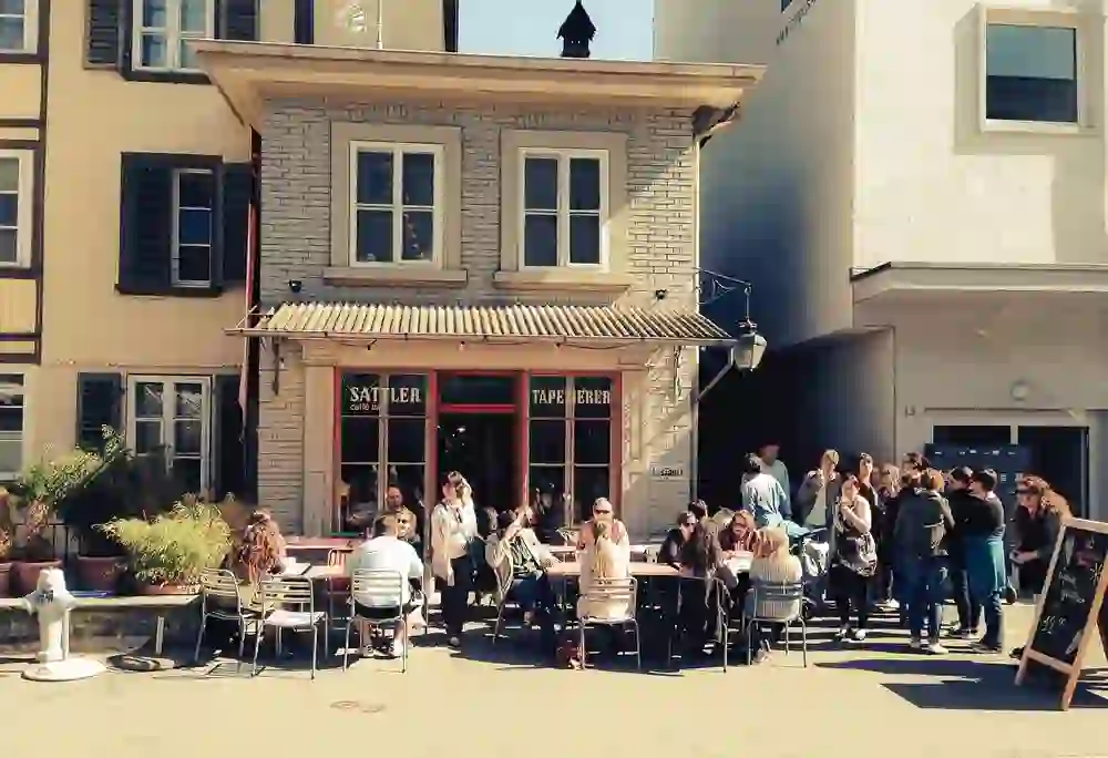 The picture shows a lively outdoor café, with guests relaxing at tables under the sun's rays. The warm and welcoming atmosphere is emphasised by the smiles and conviviality of the people. The scene conveys a sense of community and leisure enjoyment.