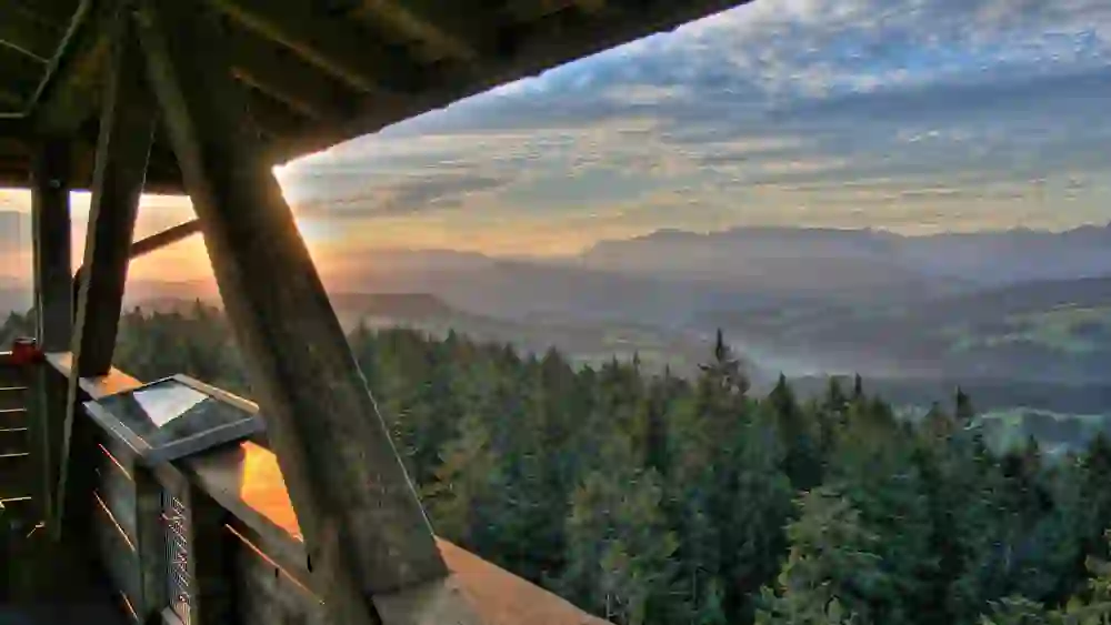 L'image offre une magnifique perspective depuis la tour d'observation Chuderhüsi à Röthenbach, caractérisée par sa balustrade naturelle en bois et sa structure de toit. La vue s'ouvre sur un vaste panorama de douces collines et de forêts qui baignent dans la lumière chaude du coucher de soleil. Le ciel est dramatique avec des motifs de nuages et des couleurs éclatantes, et au loin se dessinent les contours des montagnes, ce qui met en valeur la beauté majestueuse de la nature.