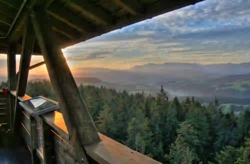 The picture offers a wonderful perspective from the Chuderhüsi viewing tower in Röthenbach, characterised by the natural wooden railings and roof construction. The view opens onto a wide panorama of rolling hills and forests bathed in the warm light of sunset. The sky is dramatic with cloud patterns and radiant colours, and in the distance the outlines of the mountains stand out, highlighting the majestic beauty of nature.
