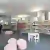 L'immagine mostra un angolo per bambini in una biblioteca. Ci sono diversi scaffali pieni di libri colorati per bambini. Al centro della stanza ci sono dei tavolini e delle sedie bianche e delle poltrone rosa. Un bambino è seduto e legge un libro, mentre sullo sfondo si vedono altri bambini e adulti. La stanza è luminosa e accogliente, con pavimenti in moquette e un'ampia illuminazione. Sullo sfondo si vede una porta di vetro, che presumibilmente conduce a un'altra area della biblioteca.