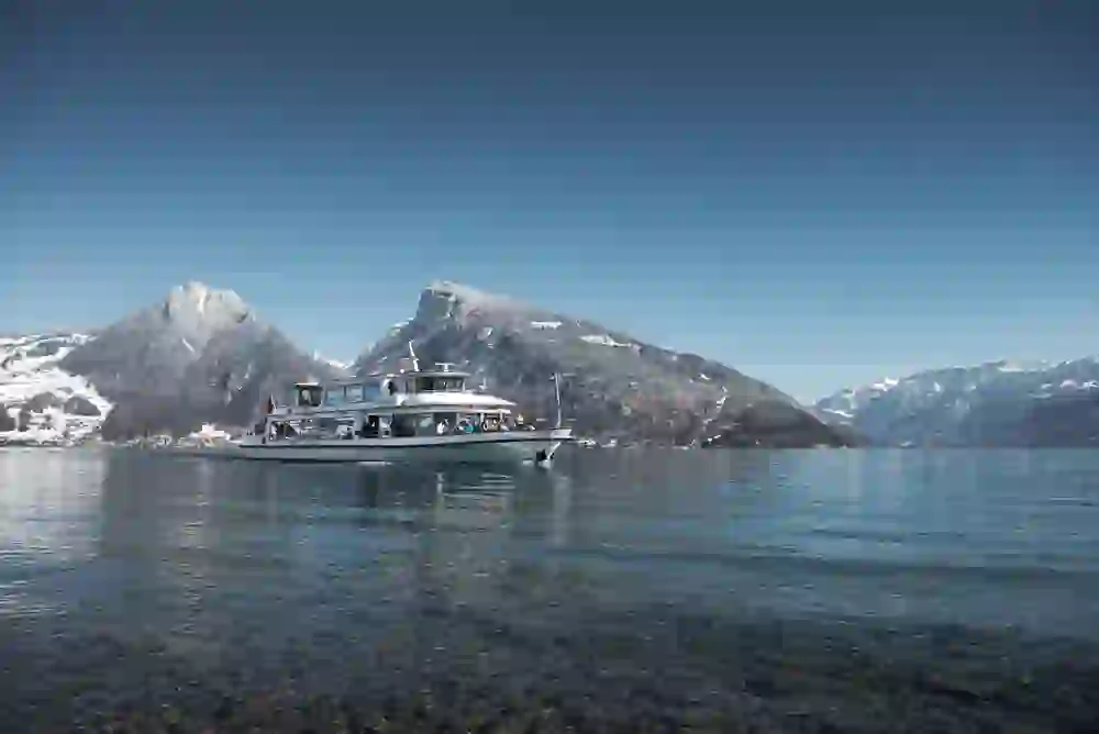 The picture presents the tranquil elegance of a winter cruise on Lake Thun. A boat glides gently over the crystal-clear water, framed by snow-capped mountains that form a breathtaking backdrop in the clear air.