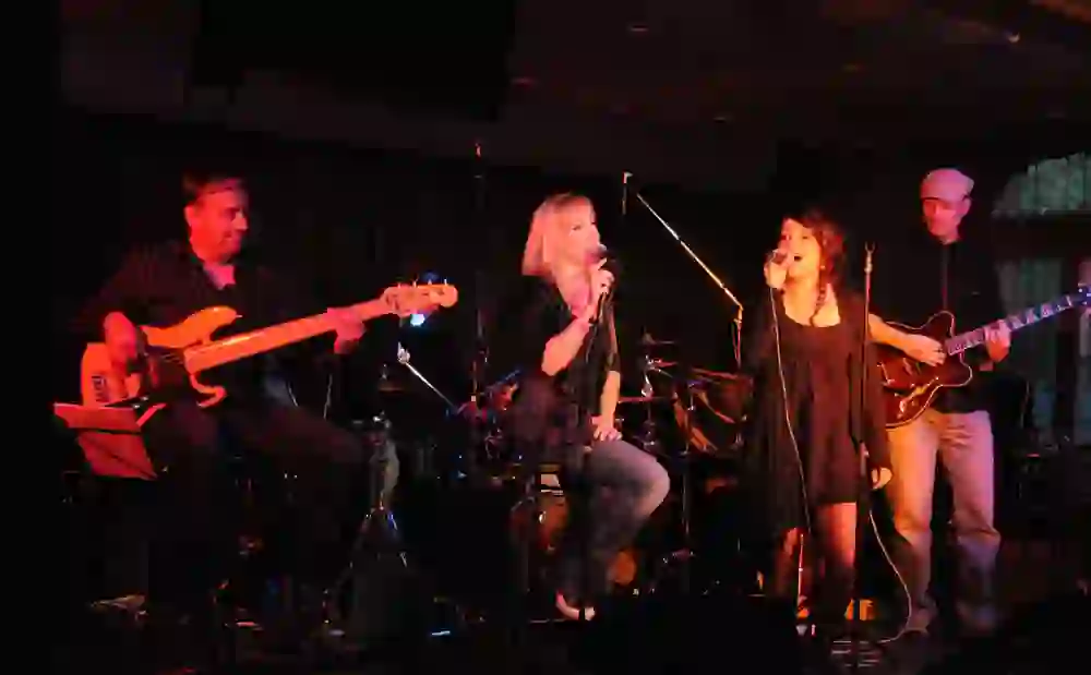 Das Bild fängt eine Band bei einem Konzert in der Mahogany Hall ein, mit Sängerinnen, die emotional performen, einem Bassisten und einem Gitarristen, die konzentriert spielen. Die intime Beleuchtung erzeugt eine herzliche und einladende Stimmung, typisch für Live-Musik-Veranstaltungen.