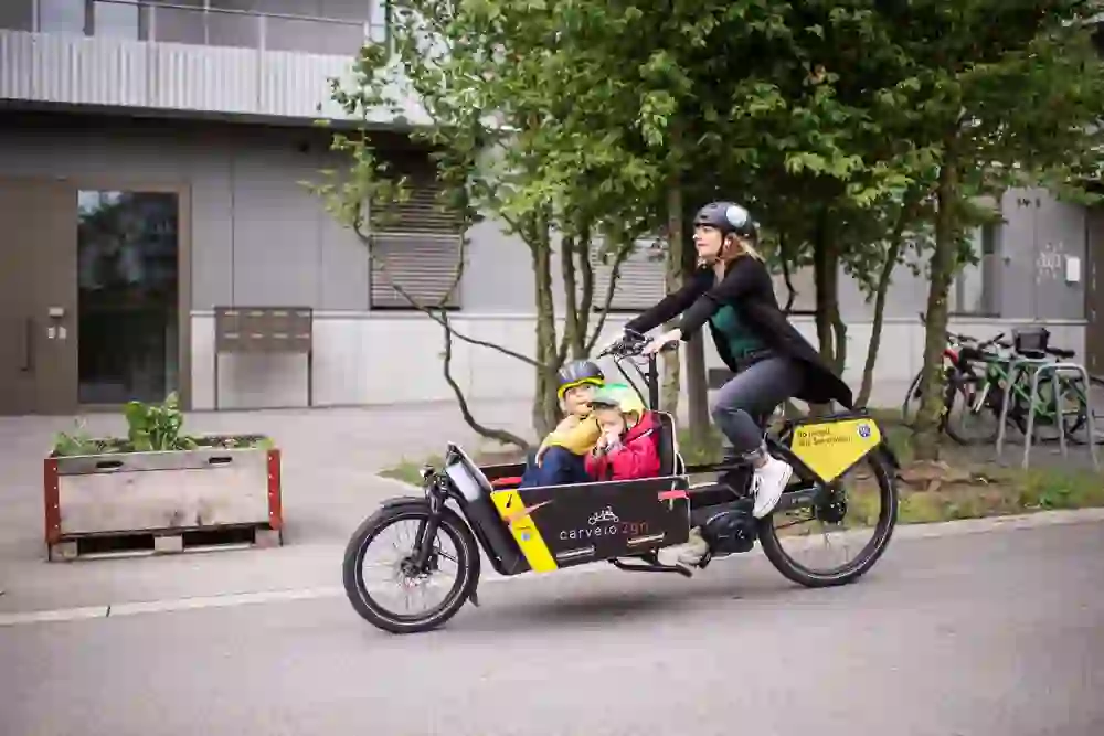 Una madre guida i suoi due figli per le strade di Berna su una cargo bike Carvelo2go. I bambini, protetti dal casco e avvolti in calde giacche, mostrano con i loro volti curiosità e gioia. Il loro viaggio urbano riflette un mezzo di trasporto sostenibile e adatto alle famiglie nella città moderna.