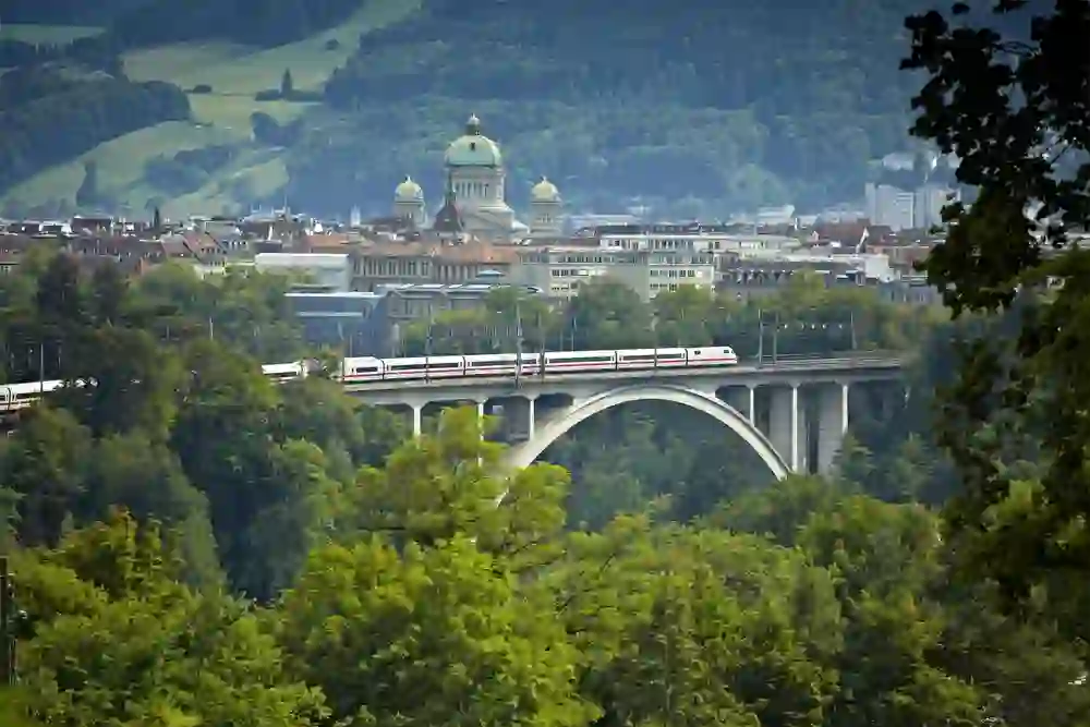 Un train traverse un pont à Berne, niché dans un écrin de verdure et derrière lui trône le Palais fédéral, une image de coexistence pacifique entre nature et architecture.