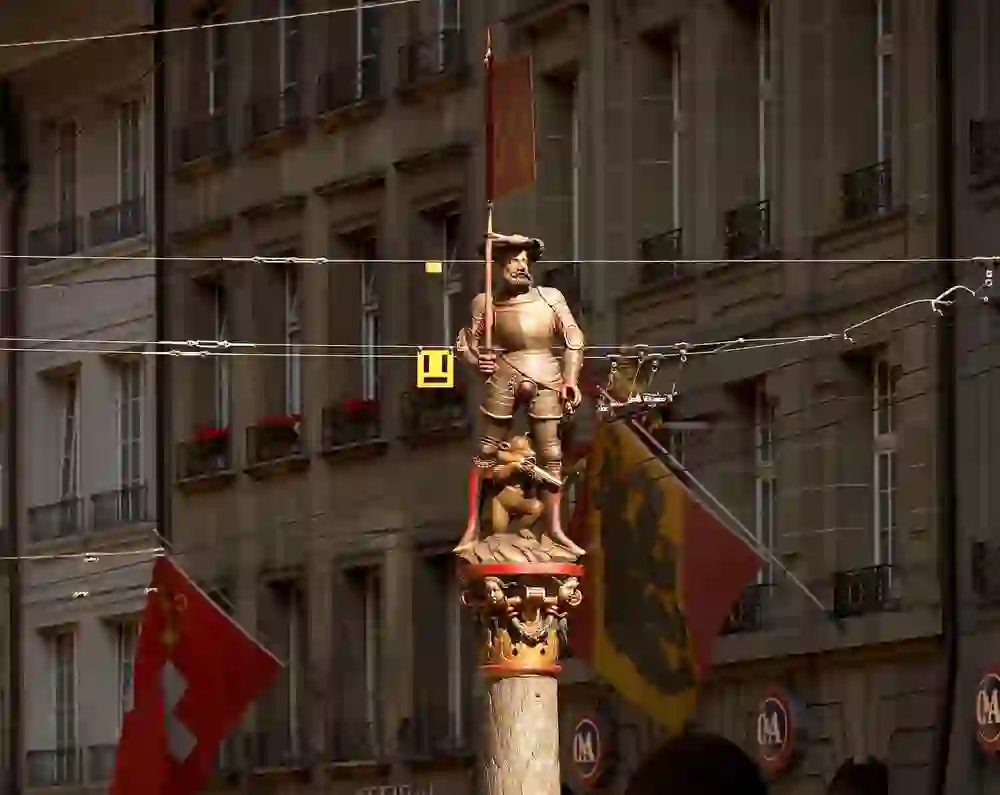 Das Foto zeigt den Schützenbrunnen in Bern, eine Statue eines Schützen in voller Rüstung, die auf einer Säule steht, vor einem Gebäude mit schweizerischen und bernischen Fahnen.