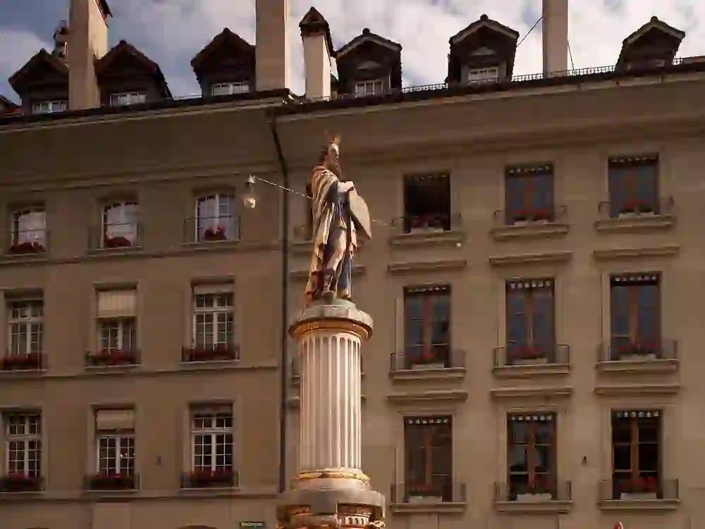 The photo shows the Moses Fountain on Münsterplatz in Bern. A statue of Moses with the tablets of the law stands on a column against the backdrop of a classical building with flower-decorated windows.