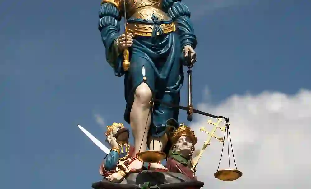 L'image montre un gros plan d'une sculpture colorée d'une Justice aveugle avec une balance et une épée, faisant partie de la fontaine de la justice à Berne.