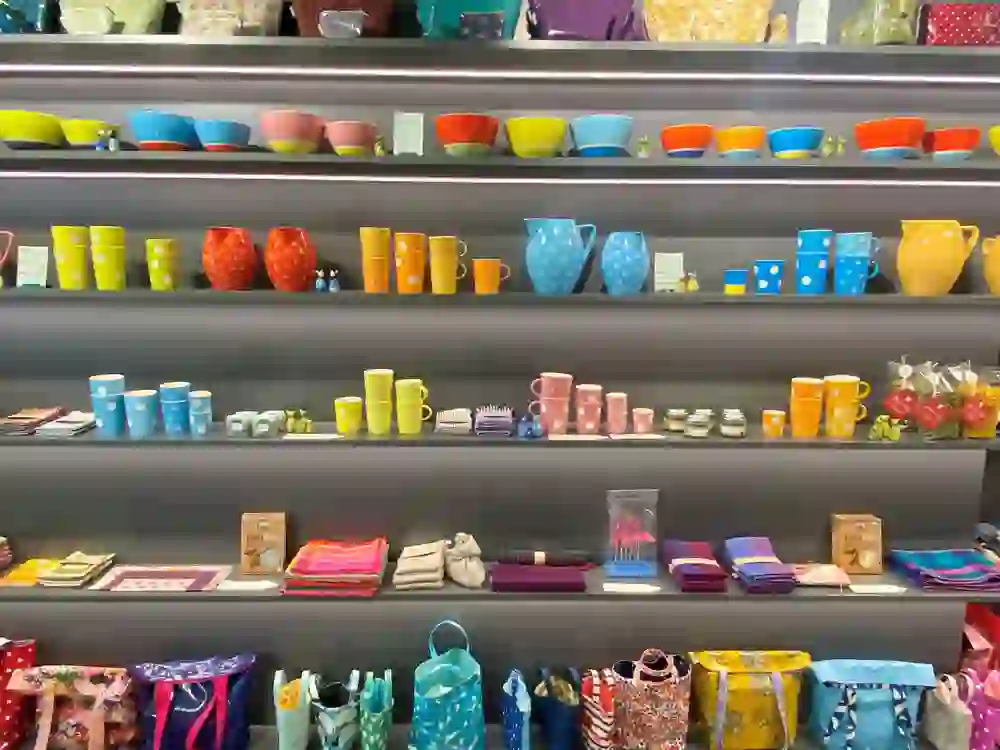 Das Bild präsentiert ein buntes Regal im "EinLaden" in Bern, das mit einer Vielzahl von farbenfrohen Geschirr und Wohnaccessoires bestückt ist. Die lebendigen Farben und Muster der Artikel strahlen Fröhlichkeit und Wärme aus, was eine einladende, heimelige Einkaufsatmosphäre schafft.