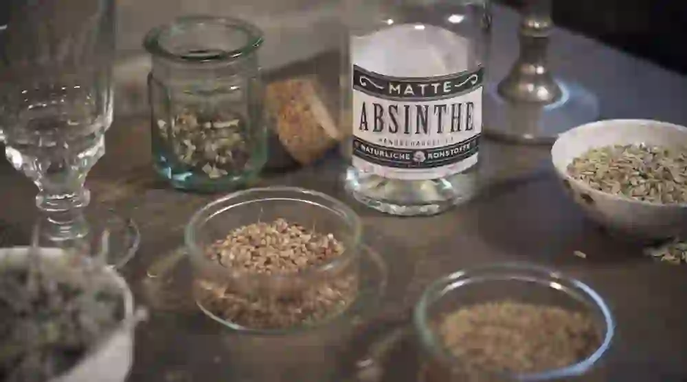 A quiet moment in the Matte distillery, captured through the interplay of traditional craftsmanship and the purity of the ingredients. An open glass of hand-crafted absinthe stands in the centre, surrounded by a collection of aromatic herbs and spices that highlight the essence of the distillation process.