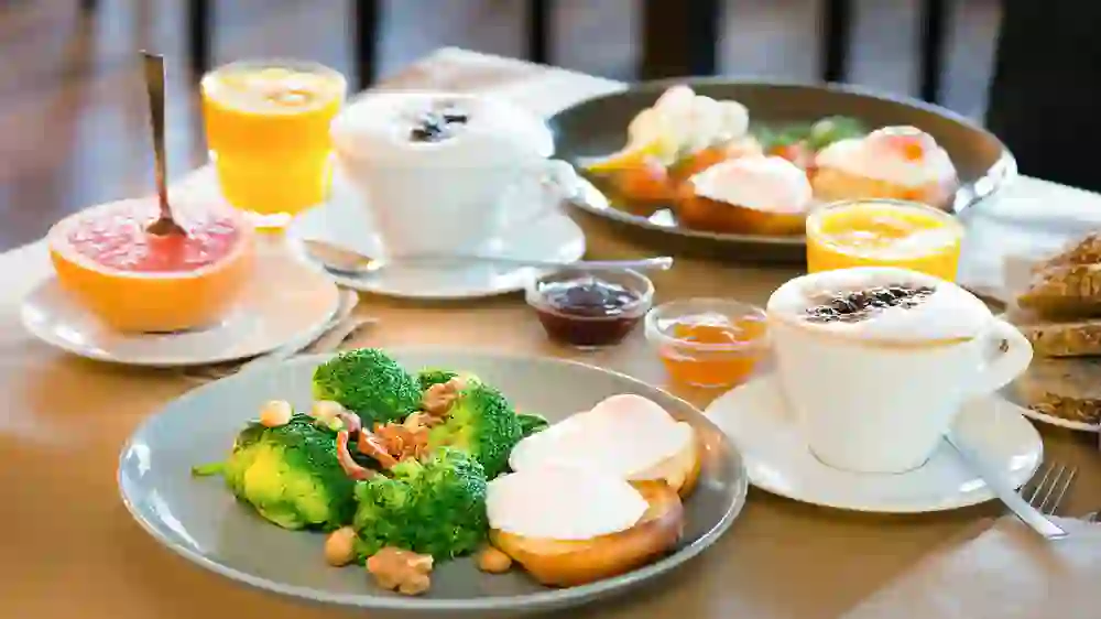 Das Bild zeigt ein Frühstücksarrangement mit gesunden Optionen. Es gibt pochierte Eier auf Toast, eine Schale mit Brokkoli und Nüssen, einen halben Grapefruit, zwei Cappuccinos, Gläser mit Orangensaft sowie Schälchen mit Marmelade und Honig. Die Präsentation ist hell und einladend.