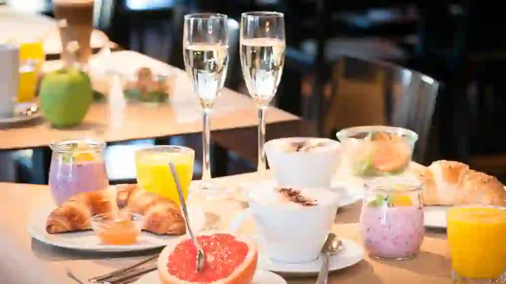 Das Foto zeigt ein Frühstück mit zwei Gläsern Sekt, zwei Croissants mit einem Becher Honig, frischem Grapefruit-Halbmond, zwei Cappuccinos, zwei Gläsern Orangensaft und zwei Joghurtbechern mit Früchten. Es wirkt einladend und festlich.