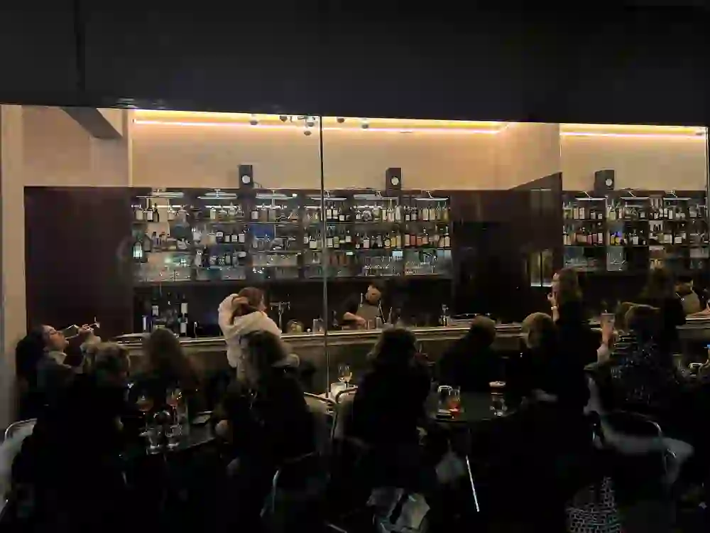 Das Bild zeigt eine belebte Bar bei Nacht mit einer gut bestückten Bartheke und mehreren Gruppen, die sich bei gedämpftem Licht unterhalten, was eine intime und gesellige Atmosphäre schafft.