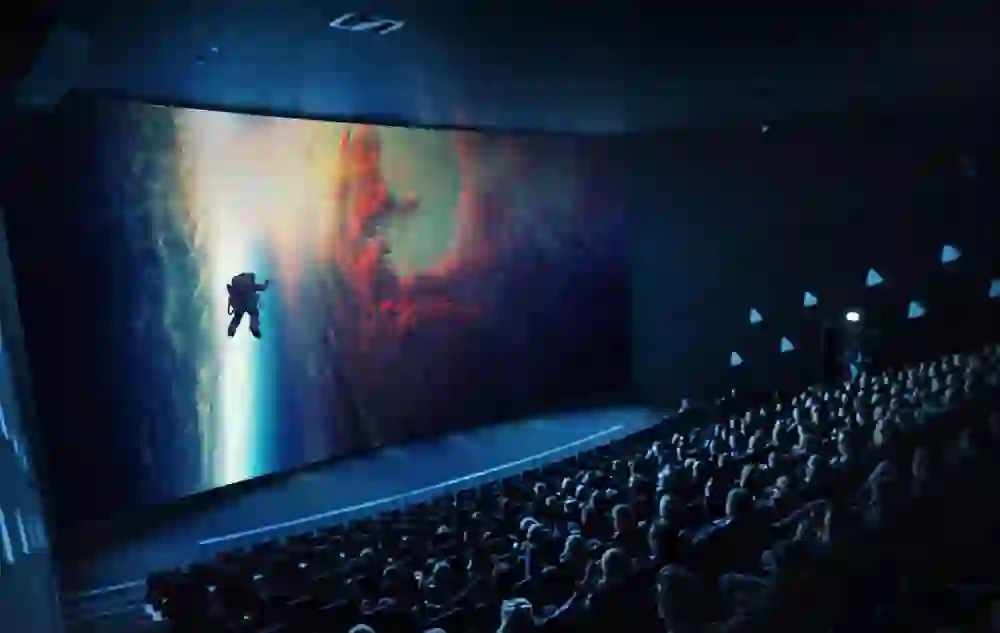 The picture shows an IMAX cinema auditorium with a stunning space film on a large screen. An astronaut floats in front of a breathtaking planet and glowing clouds. The atmosphere is impressive and immersive.