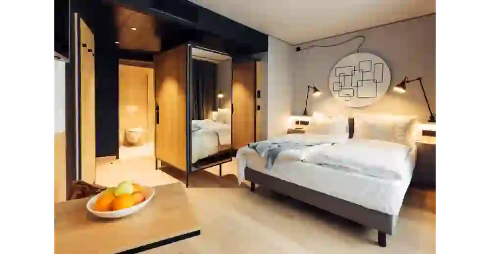 The picture shows a stylishly furnished bedroom in Harry's Home with a large, cosy bed in subtle shades of white and grey. The clean lines of the room are complemented by warm wooden elements on the wardrobes and doors, creating a pleasant balance between modernity and cosiness. A large mirror on the wardrobe door reflects the light and visually enlarges the room. An elegant, minimalist bathroom can be seen in the background. A plate of fresh fruit on the table adds a friendly, inviting accent. The room exudes comfort and stylish calm, ideal for relaxation.