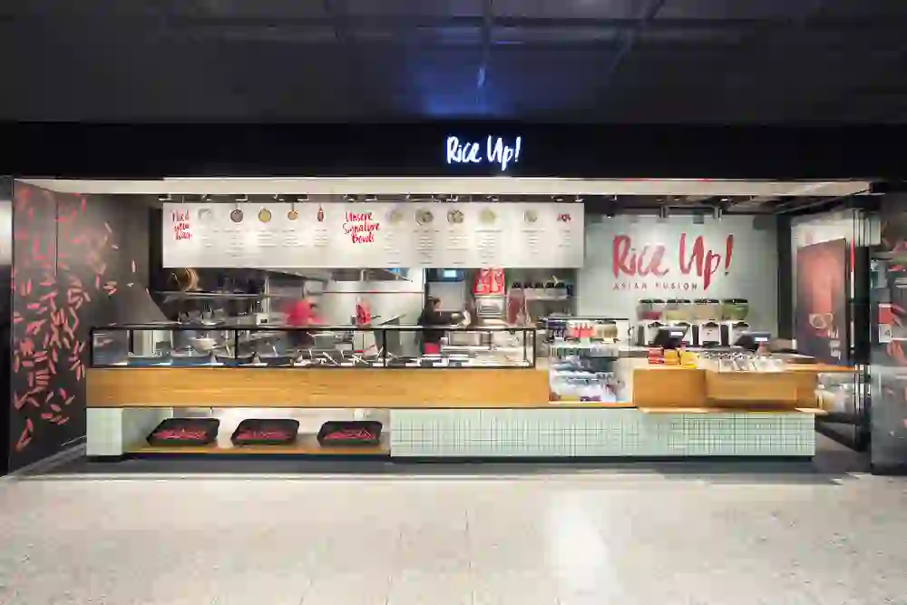 The picture shows the lively interior of the "Rice Up!" restaurant at Bern railway station. With inviting neon lighting that emphasises the logo and a warm atmosphere, the open, modern dining area invites you to linger. The staff carefully prepare dishes, while the surroundings convey an atmosphere of bustling hospitality and Asian flair.