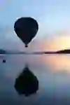 A hot-air balloon floats silently over a calm lake in the evening light. The soft pastel colours of the sky are reflected in the water and create a peaceful, dreamy atmosphere - a moment full of vastness and quiet magic.