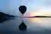 A hot-air balloon floats silently over a calm lake in the evening light. The soft pastel colours of the sky are reflected in the water and create a peaceful, dreamy atmosphere - a moment full of vastness and quiet magic.