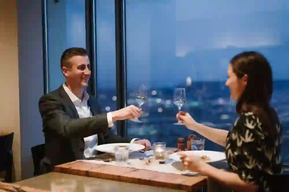 Ein Paar geniesst ein stilvolles Abendessen im Gurtners auf dem Gurten, prosten sich bei Kerzenschein zu, umgeben von der faszinierenden Aussicht auf die Lichter der Stadt durch die Panoramafenster, in einer Atmosphäre voller Wärme und Intimität.