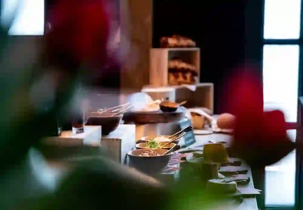 Das Bild zeigt einen Ausschnitt eines Brunch-Buffets. Man erkennt verschiedene Schalen und Platten mit Speisen, darunter Müsli und Aufschnitt, sowie ein Arrangement von Käsestücken. Im Hintergrund sind gestapelte Backwaren sichtbar, was auf eine reichhaltige Auswahl hinweist. Die Atmosphäre wirkt einladend und gemütlich.