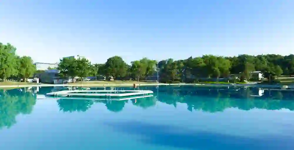 Das Freibad Weyermannshaus in Bern erstrahlt in morgendlicher Stille. Die spiegelglatte Wasseroberfläche reflektiert die umgebenden Bäume und den klaren Himmel, ein Sinnbild für Frieden und einen neuen Tag voller Möglichkeiten.