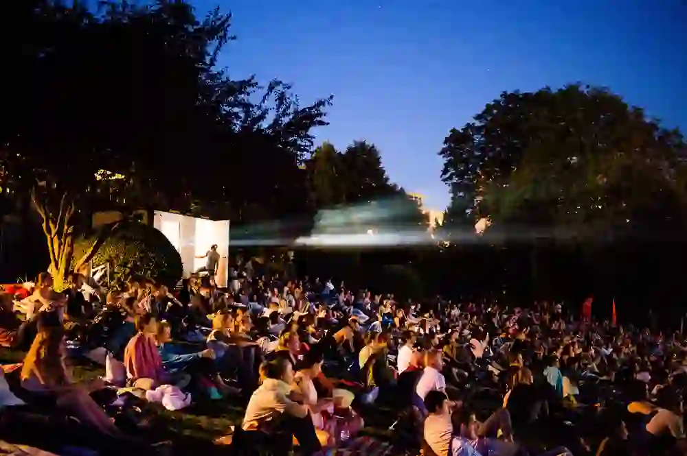 Das Bild zeigt eine große Menschenmenge, die im Openairkino Kino im Kocher unter freiem Himmel entspannt. Die Zuschauenden, verteilt auf einer grünen Rasenfläche, geniessen bei Einbruch der Dämmerung einen Film. Die Szene ist durch die Bühnenbeleuchtung atmosphärisch erleuchtet und strahlt eine gesellige und gemütliche Stimmung aus, die typisch für Sommerabende im Openairkino ist. Die Lichtstrahlen, die durch die Dunkelheit brechen, verleihen dem Ereignis ein magisches Flair.