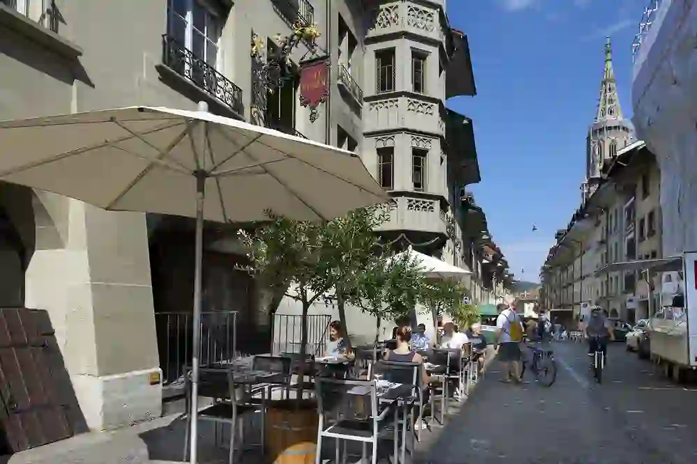 Café de rue ensoleillé dans une ruelle animée, avec des clients sous des parasols qui profitent de l'ambiance détendue de l'architecture historique.