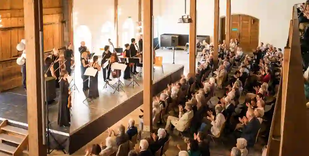 Das Bild fängt die feierliche Stimmung eines Konzertes während des Berner Kultursommers in Elfenau ein. Ein Orchester in eleganter schwarzer Kleidung spielt auf einer hellen Bühne, umgeben von einem faszinierten Publikum. Die Zuschauer, sichtlich in die Musik vertieft, applaudieren begeistert in einer historischen Halle, die ein Ambiente von Kultur und Geschichte ausstrahlt. Die warme Beleuchtung betont die Intimität und die festliche Atmosphäre dieses besonderen Moments.
