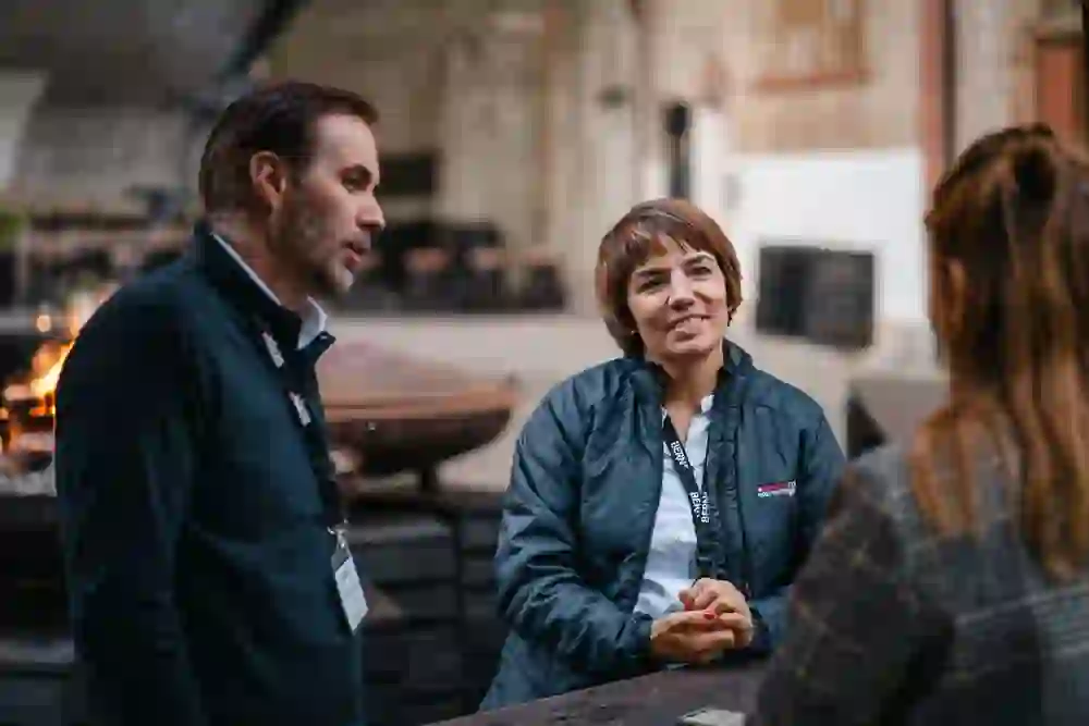 L'image montre une conversation animée entre trois personnes dans une distillerie urbaine, soulignée par l'éclairage chaleureux et l'ambiance rustique. La femme au centre sourit avec intérêt tout en écoutant attentivement, entourée du charme industriel et des flammes en arrière-plan.