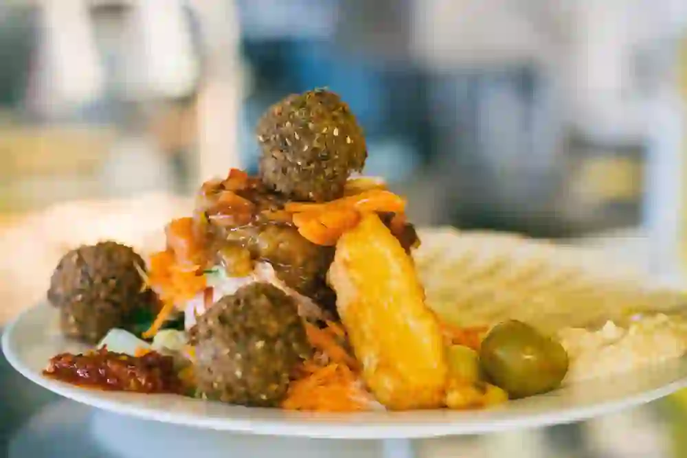 Auf dem Bild ist ein Teller mit einer appetitlichen Falafelplatte zu sehen, reichlich garniert mit frischen Gemüsen, Oliven und einer Beilage Hummus. Die knusprigen Falafelbällchen und die goldgelben Fritten laden zum Genießen ein. Die leuchtenden Farben der Zutaten und die liebevolle Präsentation erzeugen ein Gefühl von Vorfreude und kulinarischem Genuss.
