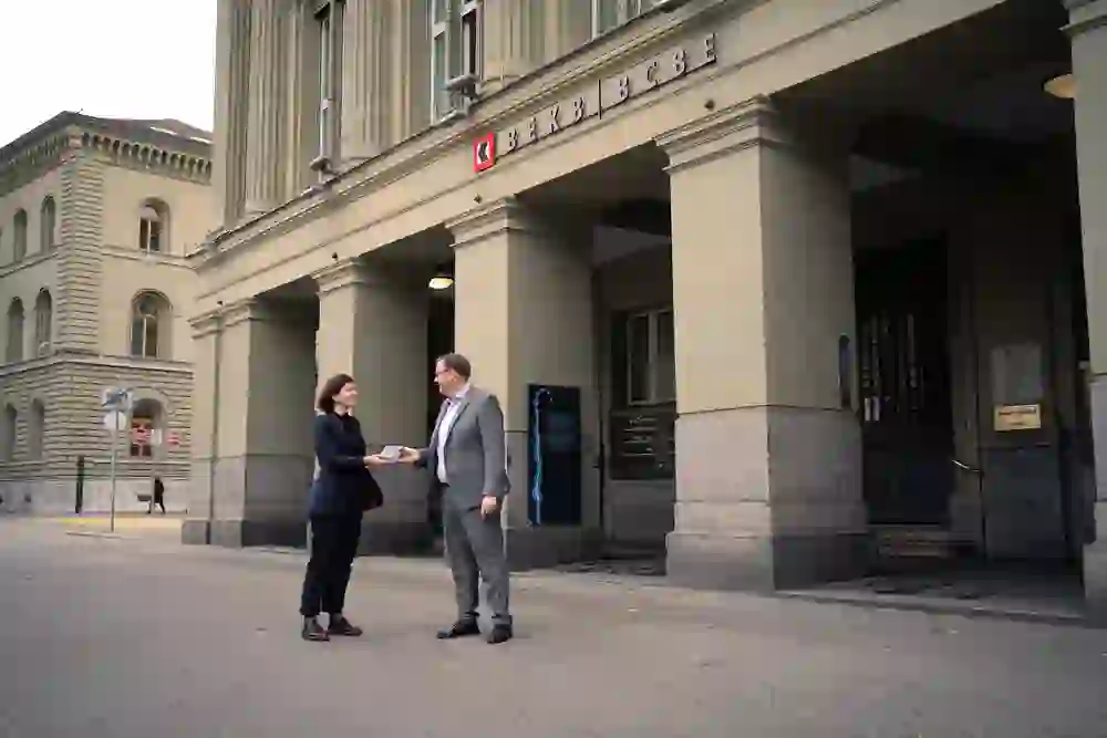 In front of a bank branch (BEKB/BCBE), a couple are engaged in a business exchange, with the woman kindly handing something to the man. The atmosphere is professional and cooperative.