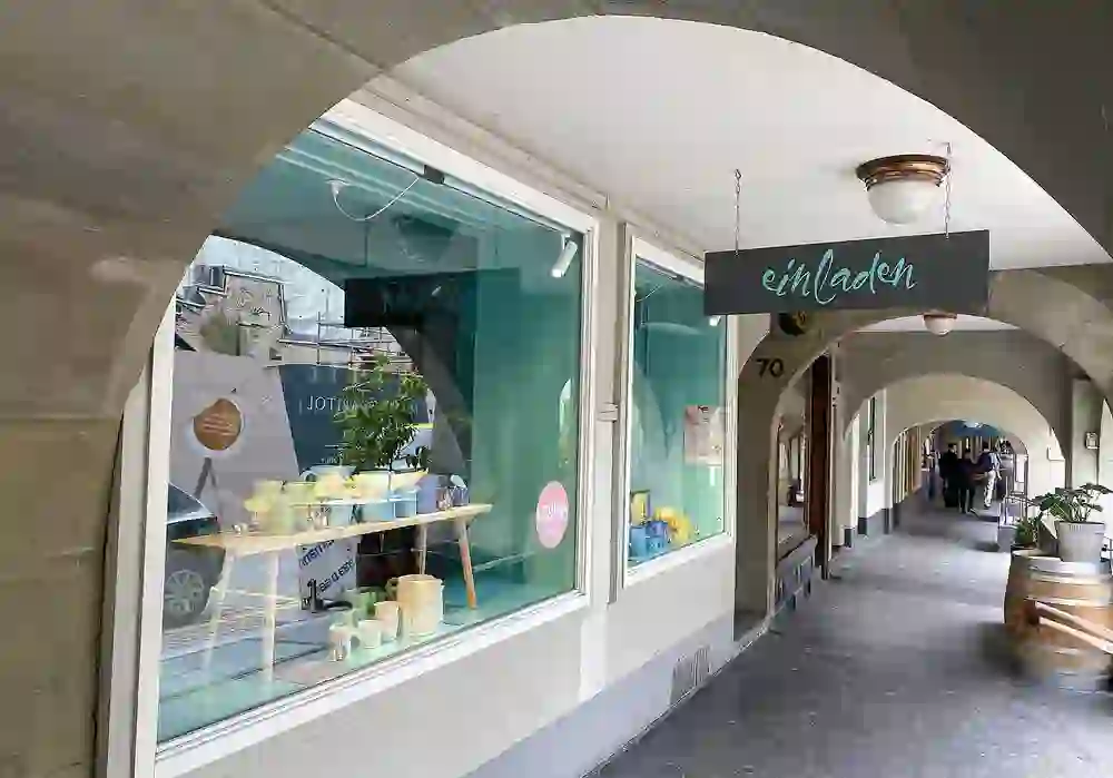 Das Bild zeigt das charmante Schaufenster von "EinLaden" in Bern, einladend unter Arkaden gelegen. Pastellfarben und fröhliche Auslagen schaffen eine heitere, freundliche Atmosphäre, die zum Stöbern und Entdecken einlädt.