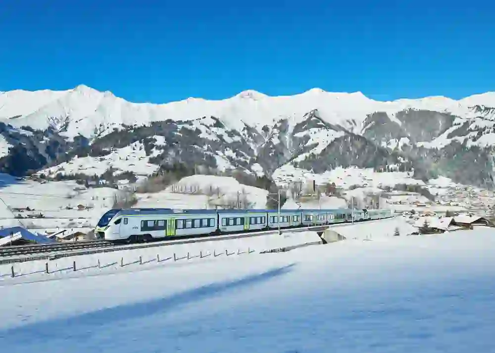 L'immagine mostra un treno moderno che attraversa un idilliaco paesaggio innevato, circondato dalle maestose Alpi svizzere sotto un cielo blu brillante.