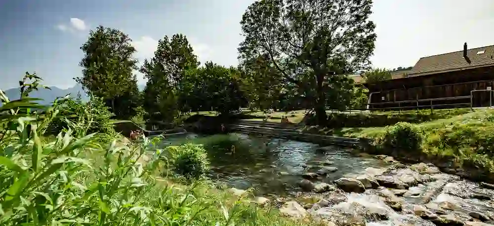 Das Bild zeigt die idyllische Gürbebadi, eine natürliche Schwimmstelle in Mühleturnen, Schweiz. Umgeben von sattem Grün und einem sanft plätschernden Fluss, geniessen Menschen einen sonnigen Tag im kühlen Nass, während im Hintergrund die ruhigen Berge emporragen.