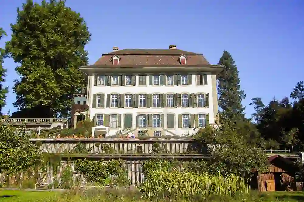 The photo shows the stately Schloss Reichenbach restaurant in Zollikofen, invitingly surrounded by a lush green landscape. The historic building with its classic shutters and characteristic hipped roof stands majestically under the blue sky. In front of the castle is a manicured garden that complements the architecture and conveys a sense of elegance and tradition. It is a place that enchants its visitors with its charm and tranquil atmosphere, perfect for a fine meal or a special event.