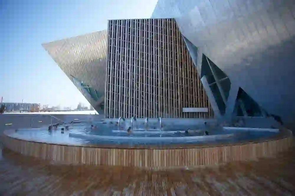 La photo montre des personnes qui se détendent dans un jacuzzi extérieur de l'espace bien-être moderne du Holiday Inn Bern Westside. L'architecture est contemporaine avec des formes géométriques marquées. L'eau claire et l'environnement recouvert de bois dégagent une impression de calme et de détente. L'atmosphère est détendue et paisible, avec une touche de luxe et de modernité.
