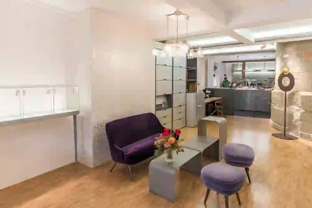 Das Bild zeigt das Innere eines eleganten Ladengeschäfts oder Showrooms, des Punctum Aureums, einem Juweliergeschäft. Es gibt eine Vitrine für Ausstellungsstücke, moderne Möbel für Kunden, darunter einen lila Samtsessel und Hocker, und ansprechende Beleuchtung. Im Hintergrund ist ein Arbeitsbereich mit Mitarbeitern zu erkennen, was auf ein Geschäft hindeutet, das kundenspezifische Arbeiten oder Reparaturen anbietet. Die Einrichtung ist modern und stilvoll, mit einer Kombination aus traditionellen und zeitgenössischen Elementen.